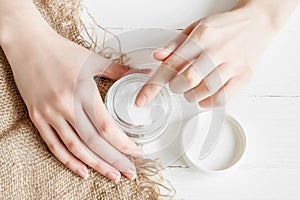 Female hands with a jar of cream on a white background. Girl applies moisturizer to her hands with dry flower petals  top view.