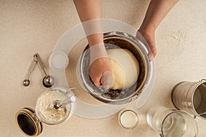 Female hands kneading dough in a bowl on tabletop. Various kitchen utensils, ingredients. Top view