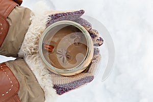 Female hands in knitted grey mittens hold white cup of spicy coffee with anisetree star.