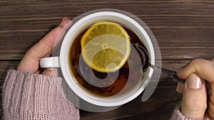 Female hands in a knitted sweater hold a cup of tea with lemon and stir it with a spoon.Close-up.