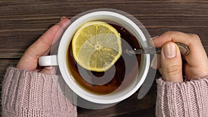Female hands in a knitted sweater hold a cup of tea with lemon and stir it with a spoon.Close-up.