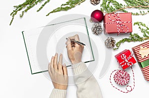 Female hands in knitted sweater are writing with pen in clean notebook plans for the new year, gift boxes, fir branches on white t