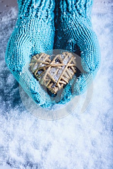 Female hands in light teal knitted mittens with entwined beige flaxen heart on a white snow background. St. Valentines Day concept