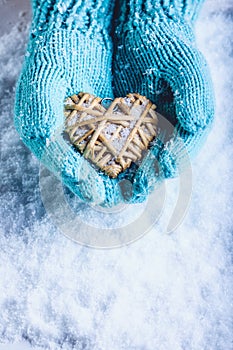 Female hands in light teal knitted mittens with entwined beige flaxen heart on a white snow background. St. Valentines Day concept