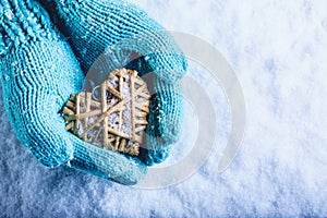 Female hands in light teal knitted mittens with entwined beige flaxen heart on a white snow background. St. Valentines Day concept