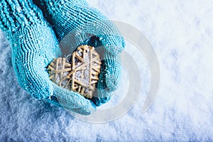 Female hands in light teal knitted mittens with entwined beige flaxen heart on a white snow background. St. Valentines Day concept