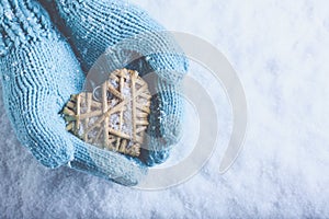 Female hands in light teal knitted mittens with entwined beige flaxen heart on a white snow. Love, St. Valentines Day concept