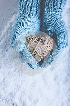 Female hands in light teal knitted mittens with entwined beige flaxen heart on a white snow. Love, St. Valentines Day concept