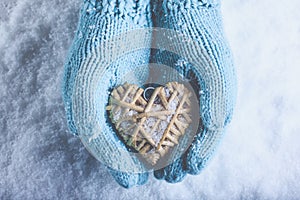 Female hands in light teal knitted mittens with entwined beige flaxen heart on a white snow. Love, St. Valentines Day concept