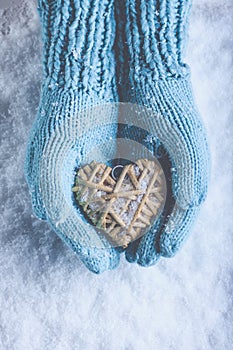 Female hands in light teal knitted mittens with entwined beige flaxen heart on a white snow. Love, St. Valentines Day concept