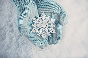 Female hands in light teal knitted mittens with sparkling wonderful snowflake on white snow background. Winter, Christmas concept