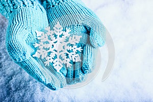 Female hands in light teal knitted mittens with sparkling wonderful snowflake on a white snow background. Winter Christmas concept