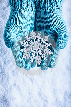 Female hands in light teal knitted mittens with sparkling wonderful snowflake on a white snow background. Winter Christmas concept