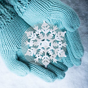 Female hands in light teal knitted mittens with sparkling wonderful snowflake on white snow background. Winter, Christmas concept
