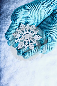 Female hands in light teal knitted mittens with sparkling wonderful snowflake on a white snow background. Winter Christmas concept