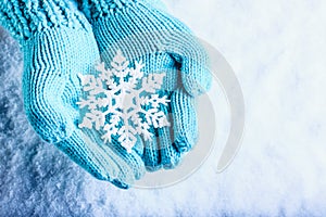 Female hands in light teal knitted mittens with sparkling wonderful snowflake on a white snow background. Winter Christmas concept