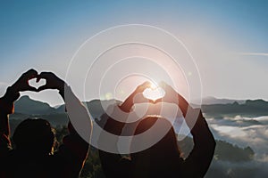 Female hands make heart shape on beautiful sunrise sky blur nature background with the sun light passing through the hands.