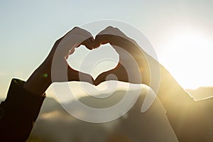 Female hands make heart shape on beautiful sunrise sky blur nature background with the sun light passing through the hands.