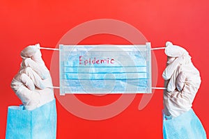 Female hands in medical gloves holding protective mask with epidemic text on it at red background. Health care concept.