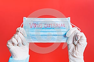 Female hands in medical gloves holding protective mask with virus outbreak word on it at red background. Health care concept.