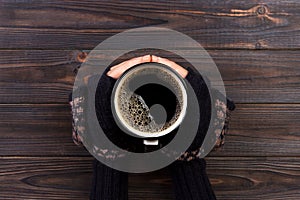 Female hands in mittens holding a cup of coffee on wooden background