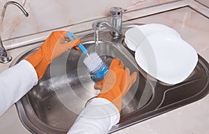 Female hands with orange gloves washing glass jar at kitchen faucet over metal sink. Close up of hand with dish brush and white