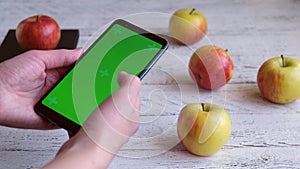 Female hands put a phone with a green screen on a wooden table with ripe apples.