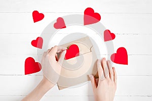 Female hands put red hearts in a congratulatory envelope on a white wooden background  happy valentine`s day background  love
