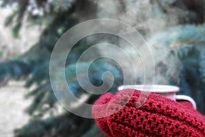 Female hands in red knitted mittens holding steaming cup of hot coffee or tea near fir christmas tree. Winter time concept.