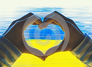 Female hands shaping heart symbol in background of yellow-blue flag of Ukraine on window. Human hands made heart shape on image of