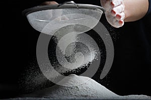 Female hands sift flour with a metal sieve on a black background.