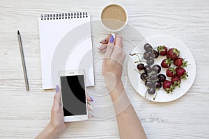 Female hands with smartphone, latte, notebook, strawberries and cherries on white wooden background, top view. Flat lay.