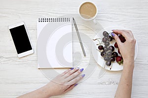 Female hands with smartphone, latte, notebook, strawberries and cherries on white wooden background, top view. Flat lay.