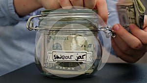 Female hands take money from Glass jar full of American currency dollars cash banknote with text SWEAR JAR. Preparation