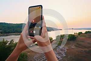 Female hands take a photo of beautiful sunset over the river on a smartphone with a broken screen.