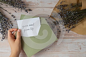 Female hands taking paper card note with text GREEN MIND GREEN LIFE from envelope. Lavender flower. Top view, flat lay