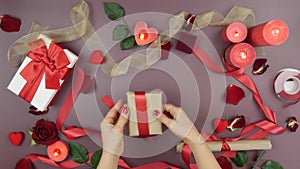 Female hands are tying red ribbon on gift box for St. Valentine`s Day on festive table background with roses, candles, gifts and h