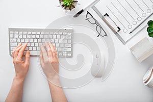 Female hands typing on keyboard at white table workplace. Home office workspace with keyboard mouse glasses. Flat lay