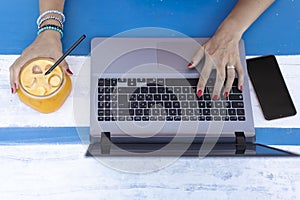 Female hands typing on laptop during remote working outdoors - Modern laptop on the blue wooden desk - Orange juice in a retro
