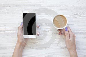 Female hands using smartphone and drinking latte on white wooden background, top view.