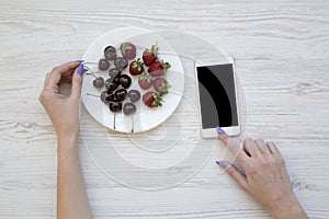 Female hands using smartphone while eating cherries and strawberries on white wooden background, top view.