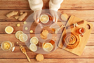 Female hands in warm sweater holding cup of tea with lemon, honey, ginger root ingredients and sweet cinnamon bun on