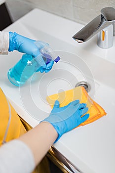 Female hands wearing rubber gloves holding microfiber cloth and spray bottle sanitizer to disinfect basin in bathroom. Protection