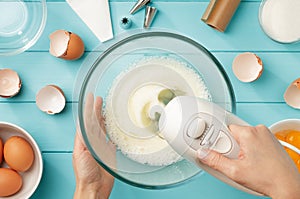 Female hands whipping egg whites cream with mixer in the glass bowl on blue pastel wooden table.
