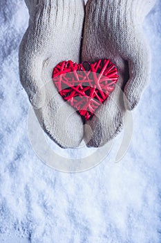Female hands in white knitted mittens with entwined vintage romantic red heart on snow background. Love and St. Valentine concept.