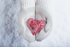 Female hands in white knitted mittens with entwined vintage romantic red heart on snow background. Love and St. Valentine concept