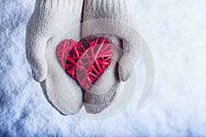 Female hands in white knitted mittens with entwined vintage romantic red heart on snow background. Love and St. Valentine concept.