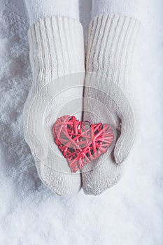 Female hands in white knitted mittens with entwined vintage romantic red heart on snow background. Love and St. Valentine concept