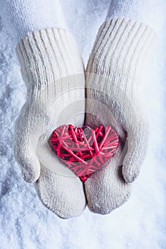 Female hands in white knitted mittens with entwined vintage romantic red heart on snow background. Love and St. Valentine concept