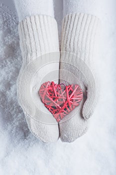 Female hands in white knitted mittens with entwined vintage romantic red heart on snow background. Love and St. Valentine concept